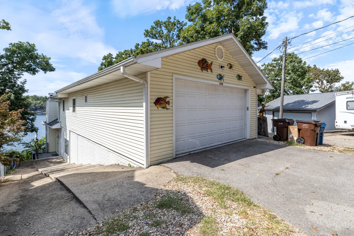 The image captures the exterior of the home with a garage door adorned with a decorative fish and a landscaped view featuring lush greenery and glimpses of the nearby lake. The driveway and open outdoor space offer a welcoming arrival point and a casual area for group outdoor gatherings.