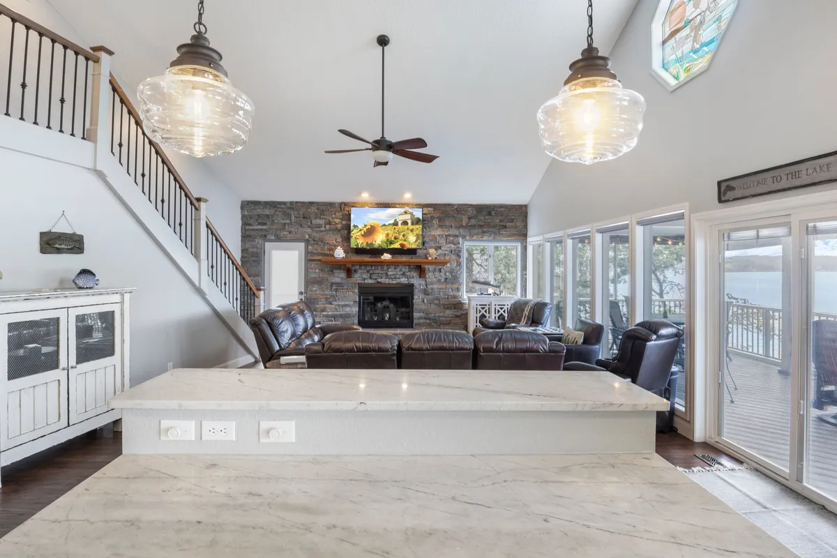 A cozy and inviting open-plan interior featuring a stone fireplace, large windows revealing outdoor views, a comfortable seating area for gatherings, and a kitchen island. The room is designed for group meals, socializing, and relaxing together after outdoor adventures.