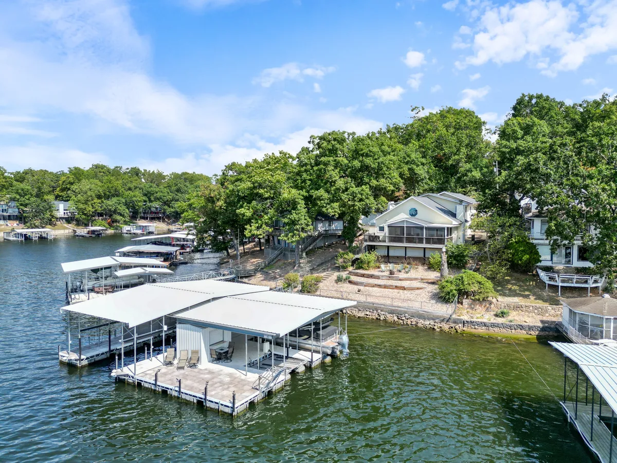 A broad exterior shot showing a lakefront property surrounded by greenery, boathouses, and a walkway leading from the house toward the water. The lake is visible, and the setting feels open, peaceful, and secure—ideal for both people and pets.