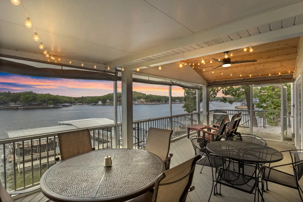 The image showcases a spacious deck overlooking the lake at sunset, featuring an outdoor dining table set under charming string lights, with comfortable seating inviting guests to relax and take in the tranquil water views.