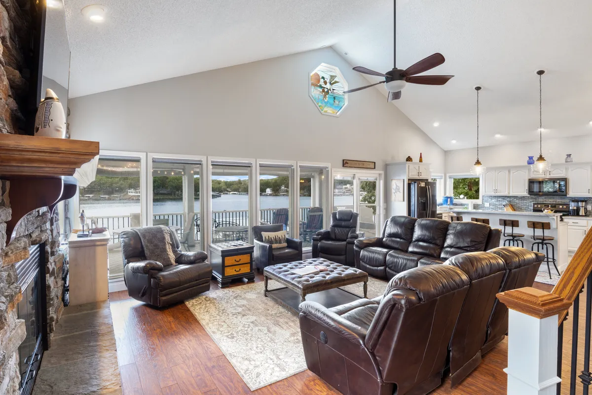 This interior shot showcases plush leather recliners facing a fireplace, with large windows offering expansive views of the lake. An octagonal window and an adjacent kitchen area add architectural interest and visual connection between spaces.