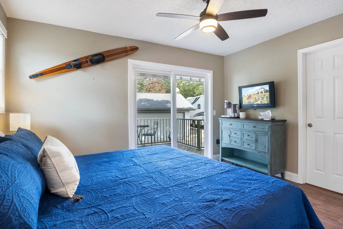 A tranquil bedroom with a blue bedspread, wooden surfboard wall decor, and a sliding glass door that allows natural light to flood the room. The space features a TV on the dresser, with soft textures and calming colors that evoke a lakeside retreat.