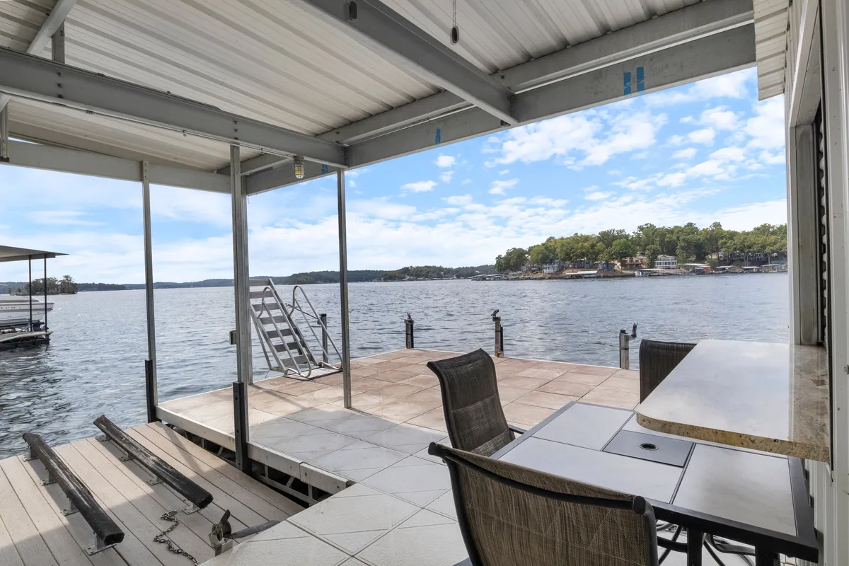 This image presents a wide view of the dock extending over the lake, with a visible staircase and outdoor dining area. The inviting setup suggests easy transitions between swimming, boating, fishing, and outdoor dining. The calm water and surrounding landscape add to the sense of relaxation and possibility for group gatherings.